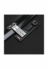 Primer plano de una maleta negra con un zip plateado y un candado de combinación. La marca dice "REDOLZ". Superficie texturizada con un diseño elegante y moderno.
