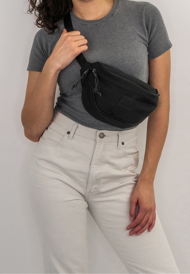Svart tyg crossbody-väska med en böjd form, utrustad med dragkedjestängning och strukturerad hårdvara. Modellen bär en grå t-shirt och vita jeans.