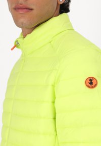 Neon yellow padded jacket with a high collar, featuring quilted sections and an orange zipper pull. Includes a round "Save the Duck" logo badge.