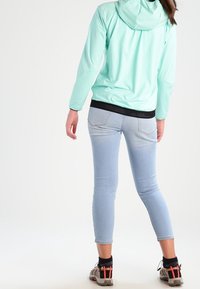 Chaqueta con capucha verde menta y cinturilla de malla negra, combinada con jeans de tobillo azul claro y zapatos de outdoor grises con acentos rojos.