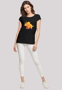 Schwarzes T-Shirt mit einem bunten, cartoonhaften Dinosaurier-Motiv; kurze Ärmel, Rundhalsausschnitt und lockerer Schnitt, kombiniert mit weißen, engen Hosen.