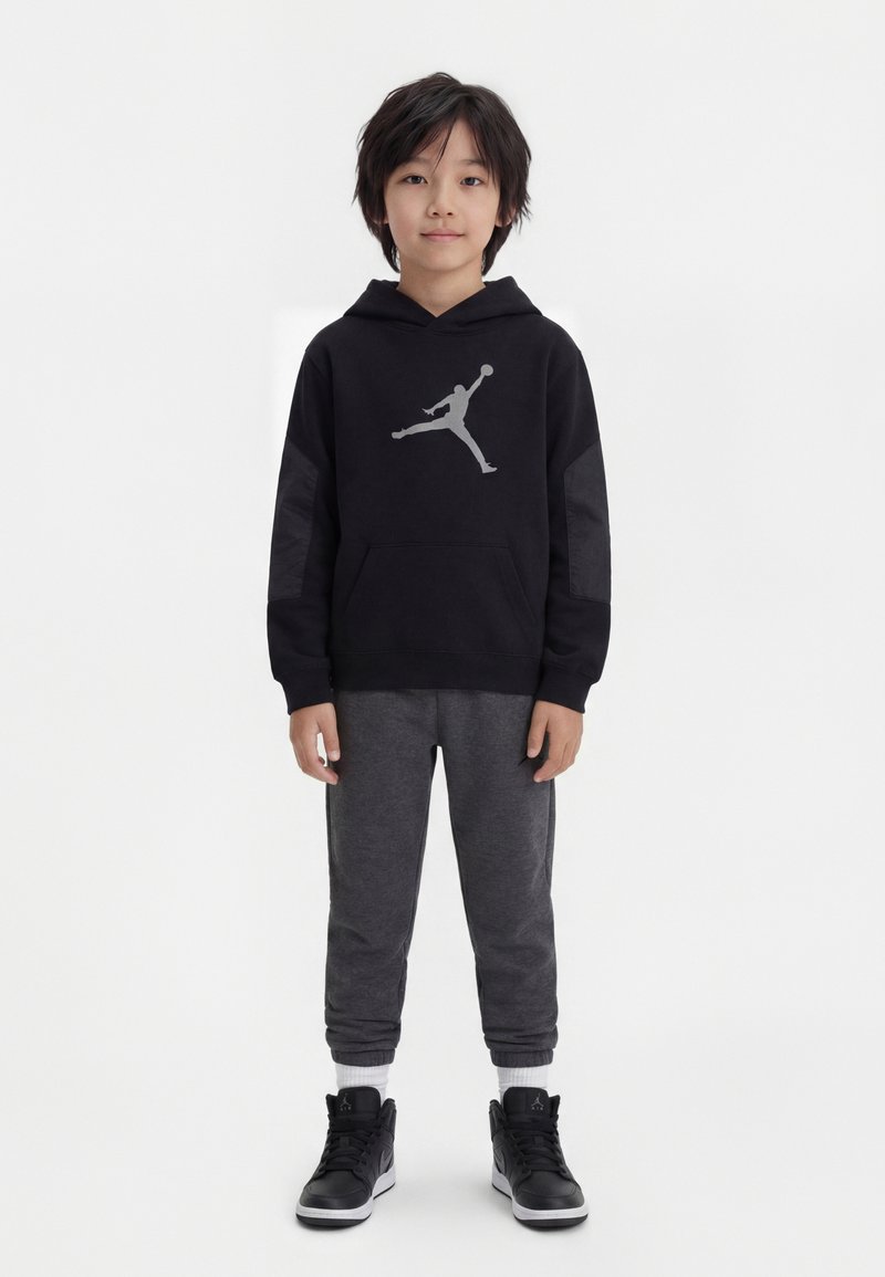 Παιδί που φοράει μαύρο φούτερ με κουκούλα με λογότυπο παίκτη μπάσκετ, σκούρο γκρι φαρδιά παντελόνια τζόγκινγκ, λευκές κάλτσες και μαύρα ψηλά αθλητικά παπούτσια, στέκεται μπροστά σε λευκό φόντο.