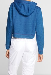 Cropped hoodie blu con cappuccio regolabile, caratterizzato da cuciture piatte e una texture morbida, abbinato a pantaloni bianchi con vita elastica.