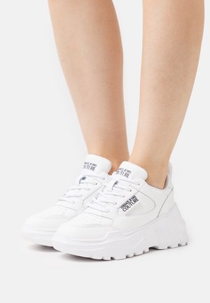 Baskets blanches à semelle épaisse, en matériau synthétique lisse, à lacets, avec un logo de marque bien visible sur le côté.