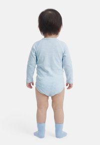 Μικρό παιδί που στέκεται με το σώμα στραμμένο προς τα πίσω, φορώντας ένα ανοιχτό μπλε φορμάκι με μακριά μανίκια και ριγέ μοτίβο, καθώς και αντιστοιχισμένα μπλε καλτσάκια, πάνω σε λευκό φόντο.