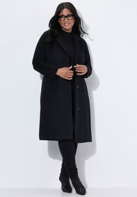 Cappotto lungo nero realizzato in un tessuto morbido, con colletto, due bottoni e una vestibilità dritta. Indossato sopra un outfit nero e stivali con tacco.