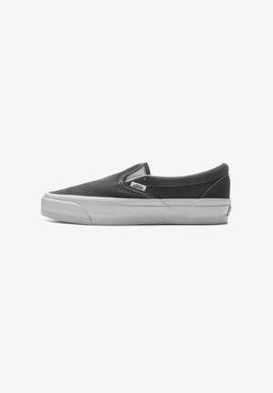 Scarpa slip-on nera in tela con punta rotonda, suola in gomma bianca e cuciture grigie. Presenta un'etichetta con il logo sul lato.