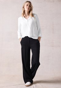 Blusa blanca de manga larga con tapeta, combinada con pantalones de pierna ancha negros y zapatillas blancas sobre un fondo claro y texturizado.