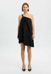 Vestido negro asimétrico con un solo hombro. Presenta tela drapeada y un detalle de nudo texturizado, combinado con tacones negros puntiagudos.