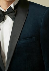 Giacca da smoking in velluto blu navy con rever a scialle in raso nero, caratterizzata da una singola tasca e una texture liscia, indossata sopra una camicia bianca.