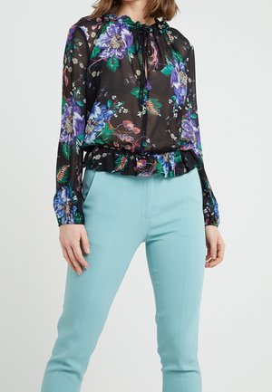 Vrouw draagt een doorschijnende zwarte bloemenblouse met paarse en groene bloemen en lichtblauwe aansluitende broek tegen een effen achtergrond.
