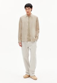 Hellebeige, vertikal gestreifte Hemdbluse, kombiniert mit einem hellbeigen T-Shirt und lockeren weißen Hosen. Die Schuhe sind weiche, tanfarbene Slipper.