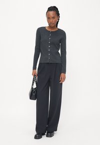 Gray button-up cardigan with long sleeves, paired with loose black trousers. Holding a small black handbag with studded accents.