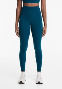 Tengerszínű, magas derekú leggings, rugalmas anyagból, sima varrásokkal és letisztult dizájnnal. Fehér sportcipőkkel párosítva.