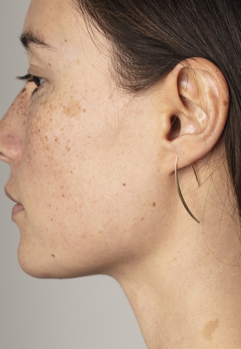 Boucle d'oreille en or avec un design incurvé, mis en valeur par une finition lisse. Affiche une esthétique minimaliste, portée sur l'oreille gauche du modèle.