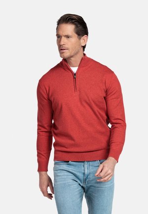 Mann trägt einen roten Pullover mit Reißverschluss bis zur Brust und hellblaue Jeans, blickt leicht nach links vor einem einfarbigen hellen Hintergrund.