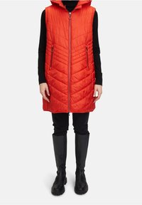 Gilet rembourré rouge avec fermeture éclair, capuche et matelassage en chevrons. Équipé de poches latérales, il est associé à un pantalon noir et des bottes hautes.