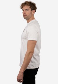 Camiseta de manga corta de color beige claro con cuello redondo y un estampado sutil del logo. La tela parece suave, ligeramente elástica y con una textura suave.