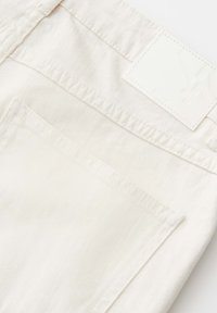 Dettaglio della tasca posteriore di jeans di denim bianco con cuciture visibili e una etichetta rettangolare in pelle vuota sulla fascia in vita.