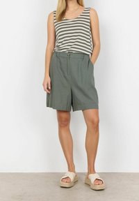 Žena v bezramenném šedobílém pruhovaném topu, zelených volných šortkách s kapsami a béžových platformových sandálech, stojící proti bílé zdi.