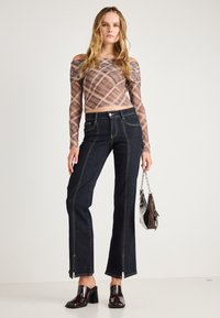 Prosojna srajca z dolgimi rokavi in karirastim vzorcem, v kombinaciji z visokimi temnimi kavbojkami z razširjenimi hlačnicami, ki imajo sprednje zadrge in kontrastno šivanje.