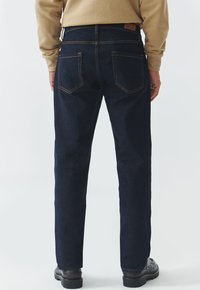 Jeans in denim blu scuro con taglio dritto, dotati di tasche posteriori e cuciture arancioni a contrasto. Tessuto resistente con una finitura liscia.