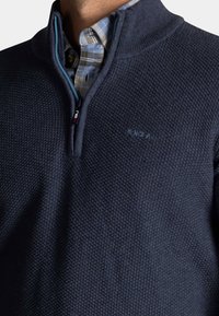 Man draagt een marineblauwe, getextureerde trui met een korte rits over een lichtblauw en beige geruit overhemd.