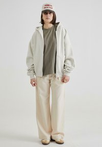 Felpa grigia chiara con zip sopra una maglietta verde oliva, abbinata a pantaloni beige a gamba larga e sneaker marroni. Cappellino bianco con scritte rosse.