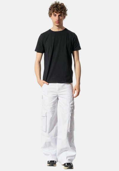 Giovane uomo con capelli ricci che indossa una t-shirt nera, pantaloni cargo bianchi e sneaker nere, in piedi con una mano in tasca.