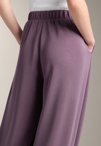 Lila weite Hosen mit glatter Textur, elastischem Bund und seitlichen Taschen, von hinten gezeigt.