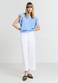 Blusa azzurra chiara con maniche arricciate, abbinata a pantaloni bianchi a gamba larga. La modella indossa scarpe leopardate nere e beige.