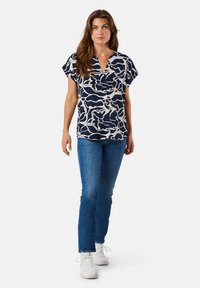 Marineblaues kurzärmeliges Blusenshirt mit weißem, welligem Muster, V-Ausschnitt-Design, kombiniert mit blauen Jeans und weißen Sneakers. Glatte Stoffoberfläche.