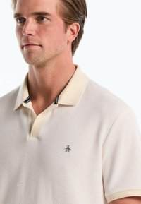 Polo-shirt in gebroken wit, van textuurstof met een kraag. Marineblauwe strepen op de kraag accentueren en een klein zwart pinguïnlogo op de borst.