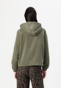 Kaki kordfløyelsjakke med hette, med ribbet tekstur og løs passform. Kombinert med brune leopardmønstrede bukser.