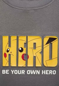 Grijze sweatshirt met grote gele letters die "HERO" spellen en een illustratie van Pikachu. Text eronder luidt: "WEES JE EIGEN HELD."