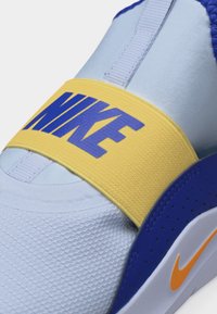 Gros plan sur une basket Nike bleu clair avec une sangle jaune portant le texte "NIKE" en bleu, et des accents bleus et orange sur la semelle et le talon.