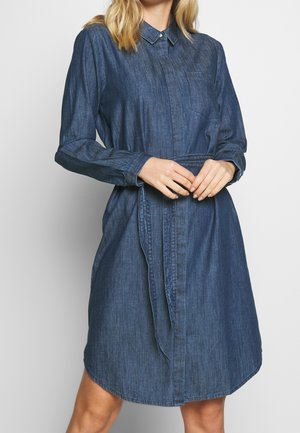 Vrouw draagt een lange mouw, knoopjes denim jurk met een kraag en tailleband, staand tegen een effen lichte achtergrond.