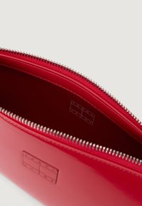 Rød glinsende pose med sølvglidelås, med en jevn tekstur og preget "TOMMY JEANS"-logo innvendig. Innvendig er den ensfarget rød.