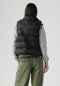 Chaleco acolchado negro, con cuello alto y diseño acolchado, llevado sobre un sudadera gris, con pantalones verdes oliva visibles por debajo.