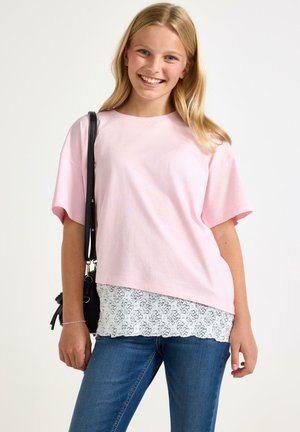 Smilende ung kvinde med blondt hår iført en lyserød skjorte over en hvid blondebluse, blå jeans og med en sort skuldertaske.