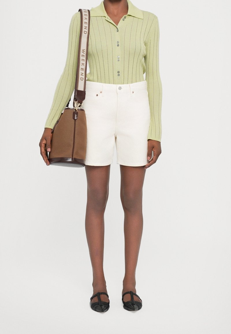 Personne portant un haut à manches longues côtelé vert clair, un short blanc taille haute, des ballerines noires pointues et portant un sac seau marron avec une large bandoulière.