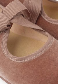 Chaussures en tissu brun avec une texture lisse, une ouverture arrondie et une sangle plate assortie en ruban. L'intérieur présente une semelle contraste claire.