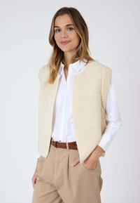 Veste en tricot beige à devant ouvert, tissu texturé et épaules larges, portée sur une chemise blanche boutonnée et un pantalon beige, avec une ceinture marron.