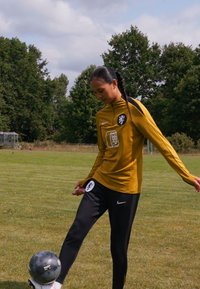 Gul atletisk langermet skjorte med svarte detaljer, svarte bukser og logoer; person kontrollerer en grå fotball på gressbane.