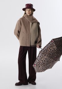 Bruine tekstuur oversized jas met een beige paneel, gecombineerd met bordeauxrode wijde broek en een luipaardprint paraplu. Bordeaux leren hoed.