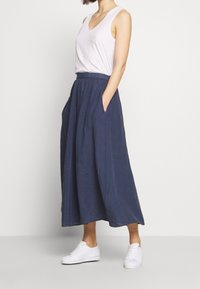 Navy blue linen maxi skirt with an elastic waistband, featuring side pockets and a flowy, A-line silhouette, paired with a light tank top and white sneakers.