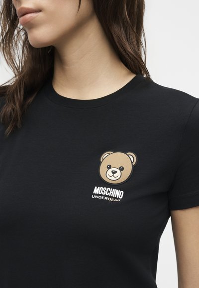 Κοντινό πλάνο ενός ατόμου που φοράει μαύρο μπλουζάκι με μικρό πρόσωπο αρκούδας και το κείμενο "Moschino Underbear" στο στήθος.