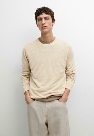 Jonge man staat met handen in zijn zakken, draagt een beige trui over een bruin overhemd en lichtgekleurde broek, tegen een effen witte achtergrond.