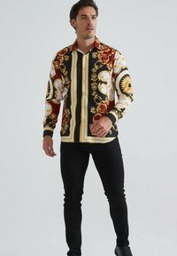 Homme debout portant une chemise à manches longues avec des motifs baroques ornés en rouge, noir, or et blanc, associée à un pantalon et des chaussures noires.
