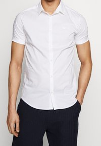 Bílá košile s krátkým rukávem a zapínáním na knoflíky, se špičatým límcem, vyrobená z hladkého materiálu. Obsahuje malé logo blízko hrudi a má přiléhavý střih.